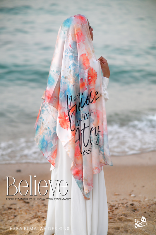 Woman wearing the Believe Silk Satin Hijab by Heba Elmalah Designs on the beach, featuring soft coral, aqua, and blush floral prints over a light background.