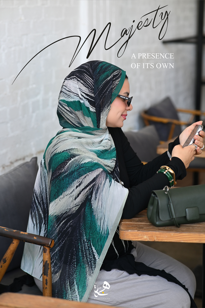 A woman wearing a stylish green, black, and white patterned hijab sits at a wooden table in a cafe, looking at her phone. The word 'Majesty' is written in elegant black script across the top, and a matching green handbag rests on the table next to her.