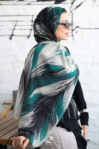 A woman wearing a teal, black, and mint green abstract-patterned hijab and sunglasses, standing in front of a white brick wall.