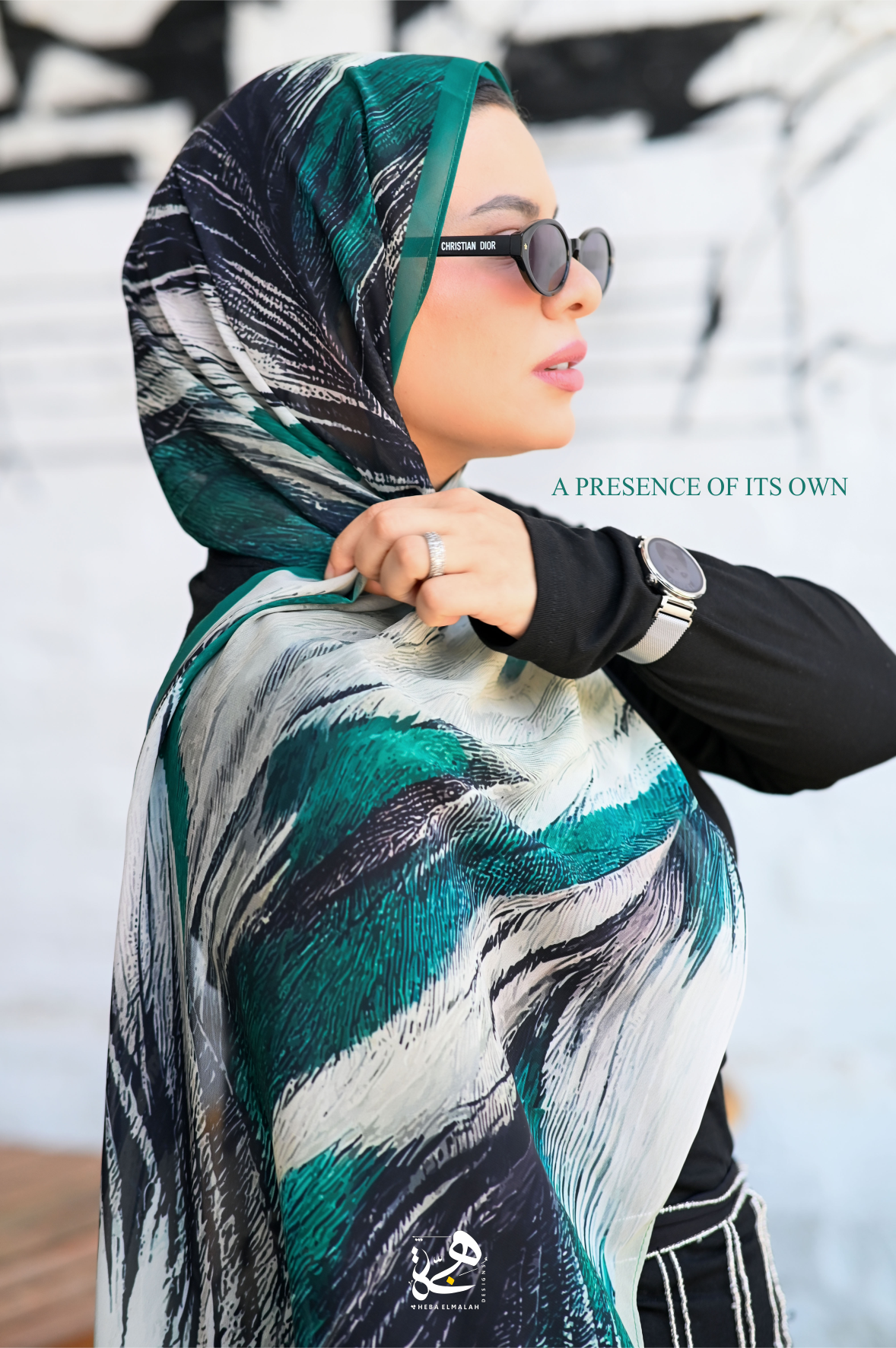 A woman wearing a patterned hijab in shades of teal, black, white, and gray, styled with a black long-sleeve top and sunglasses. The background is a white wall with black graffiti-style markings.
Luxury printed chiffon hijab Majesty